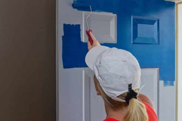 A professional painter in a white cap carefully applying a vibrant blue coat of paint to a white interior room door using a roller, provided by Siam Engineering.